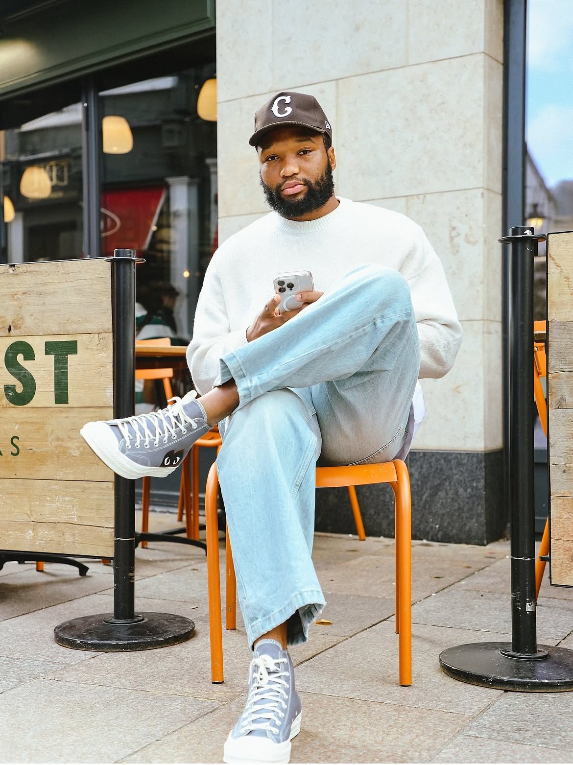 Lukhanyo Am seated outside a café in a cap, cream knit, light denim and high-tops